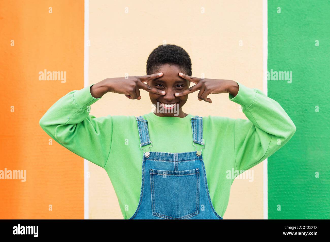 Femme heureuse montrant le geste de signe de paix et le visage touchant devant le mur multicolore Banque D'Images
