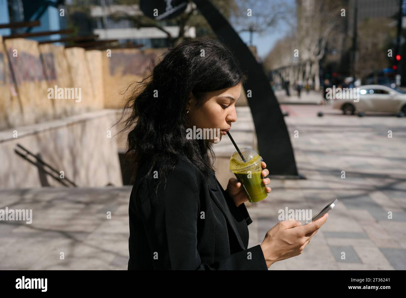 Femme d'affaires utilisant un téléphone intelligent et buvant un smoothie vert Banque D'Images