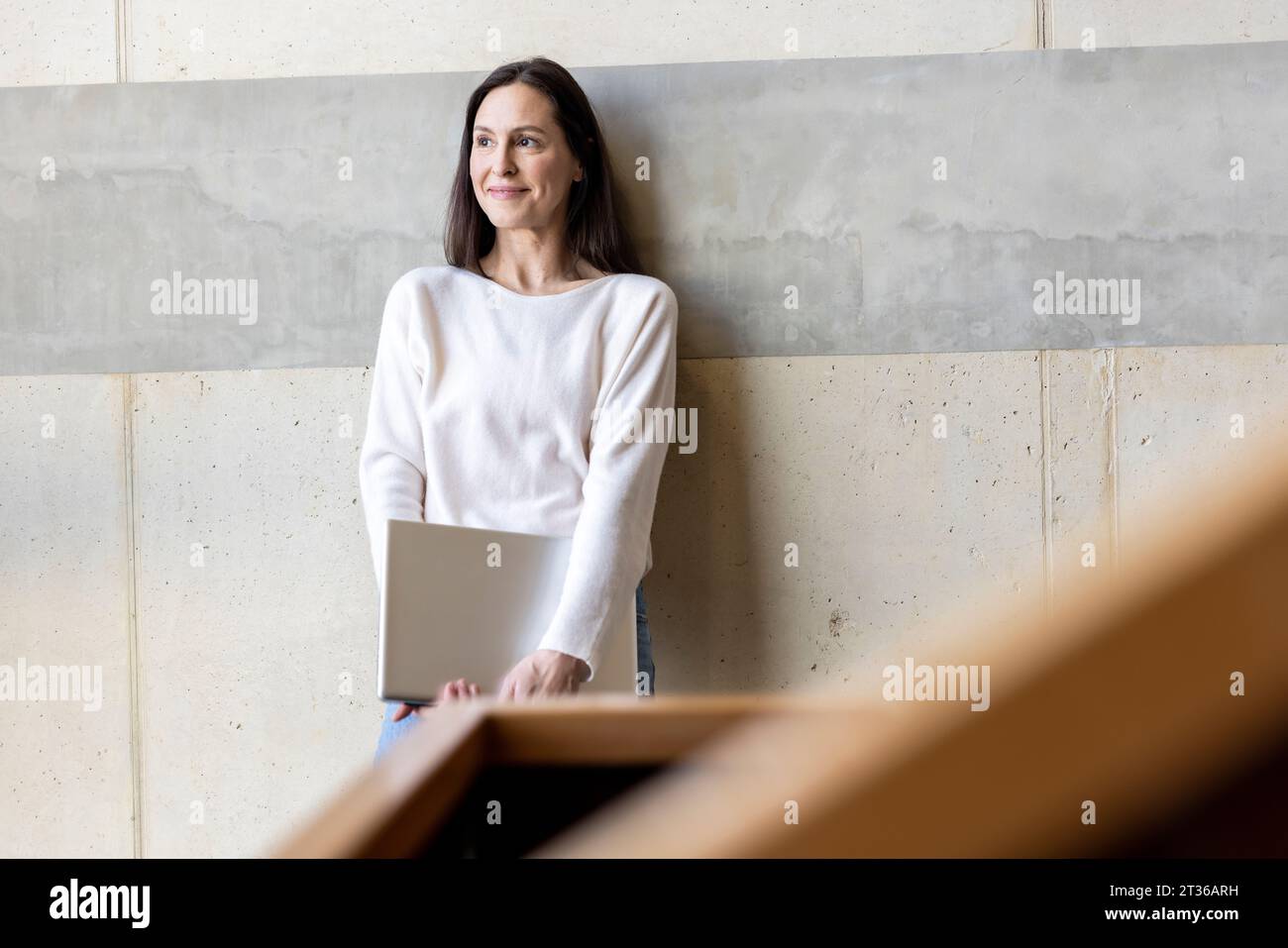 Femme d'affaires mûre réfléchie debout avec ordinateur portable devant le mur Banque D'Images