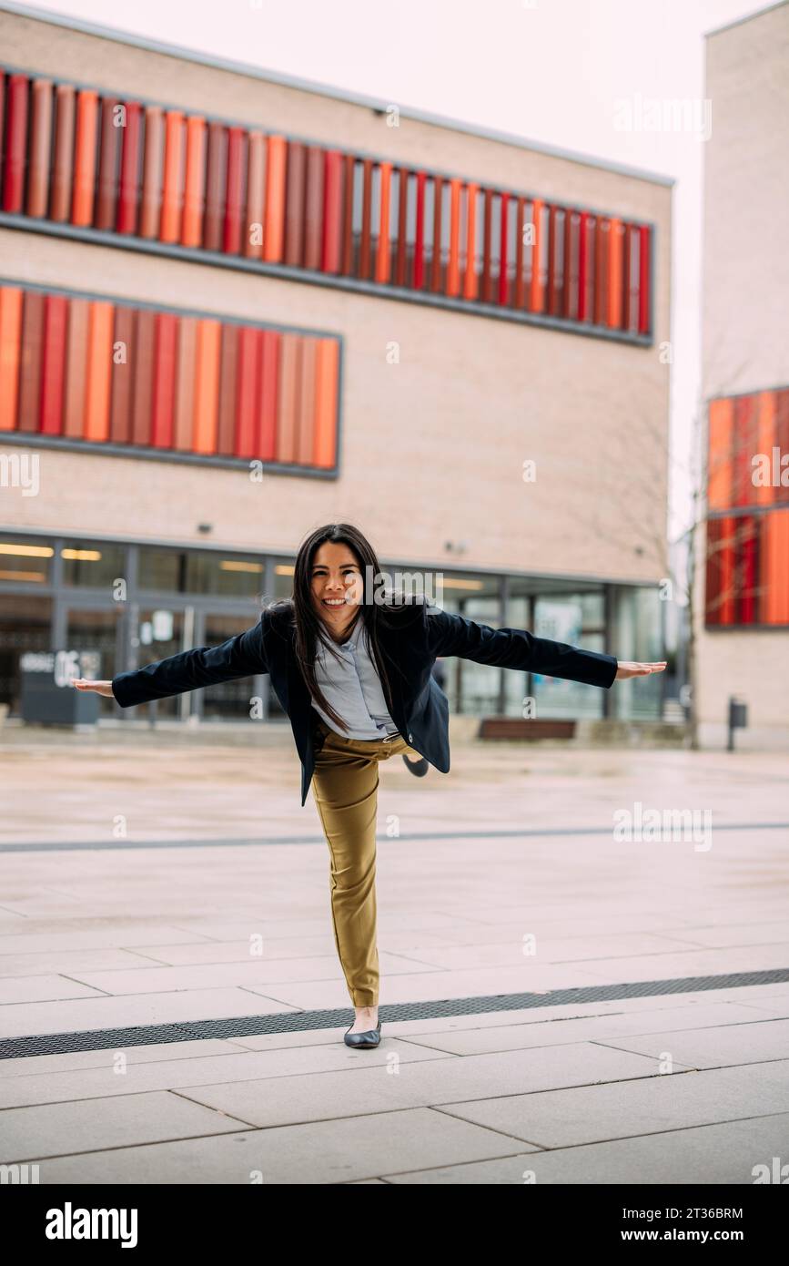 Femme d'affaires souriante debout avec les bras tendus sur une jambe Banque D'Images