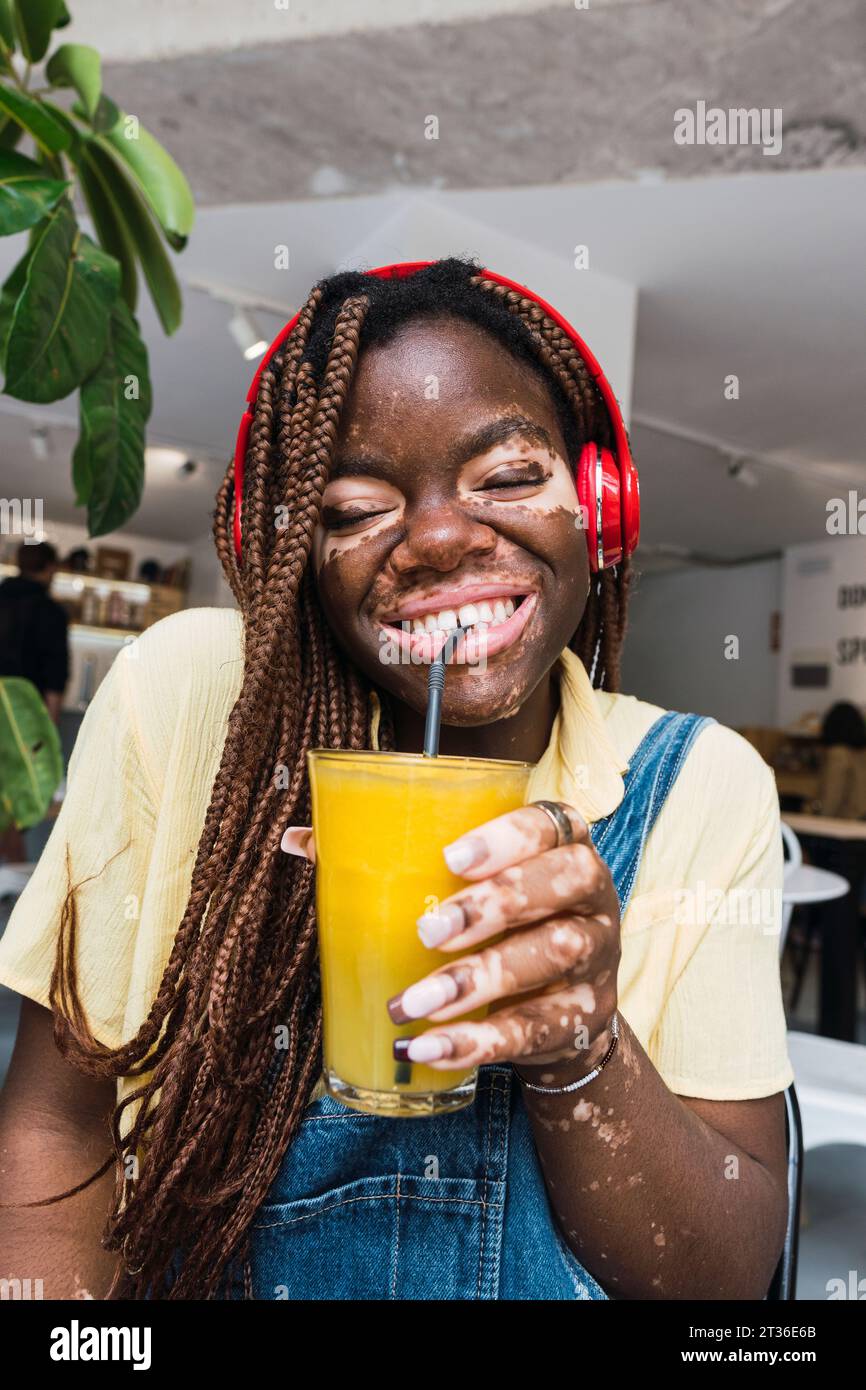 Heureuse jeune femme portant des écouteurs sans fil et buvant du jus dans le café Banque D'Images