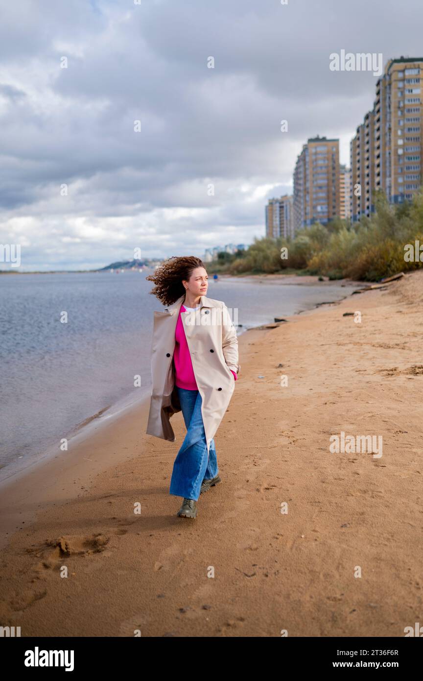 Jeune femme avec les mains dans les poches marchant sur le sable au bord de l'eau Banque D'Images