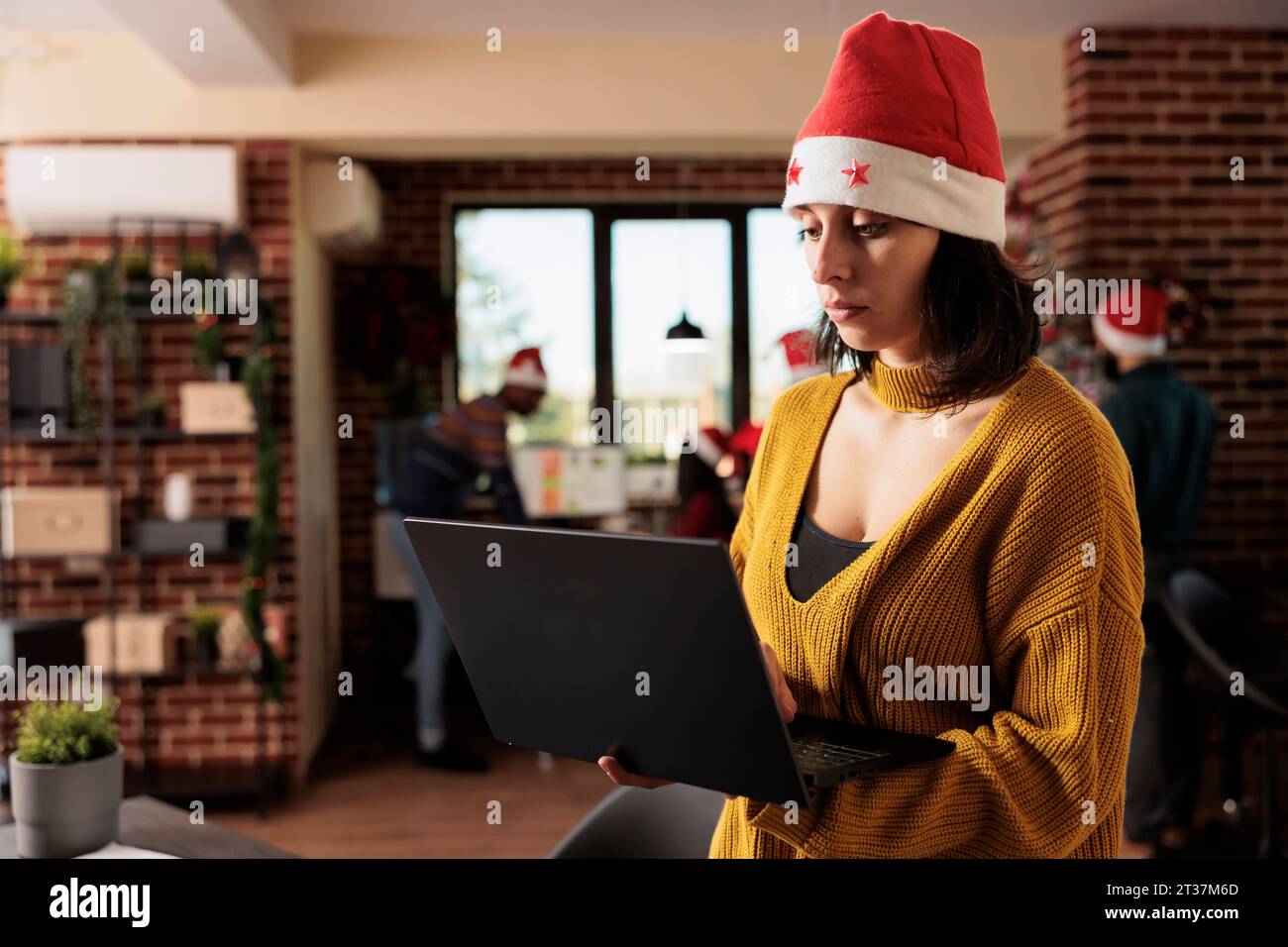 Femme chef de projet portant le chapeau de père noël gérant les tâches sur ordinateur portable tout en se tenant dans le bureau de fête d'hiver. Employé tenant un ordinateur portable et travaillant pour la date limite à la veille de noël Banque D'Images