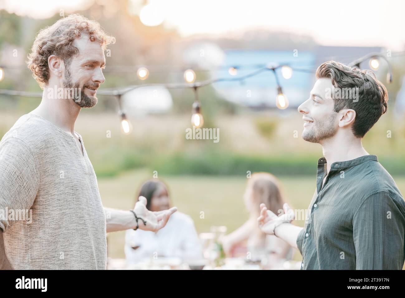 Une scène vibrante comme un groupe multi-ethnique d'amis se rassemble pour une fête de jardin. Au premier plan, deux amis masculins engagent une conversation, tandis que d'autres s'assoient à table, partagent repas et conversations, créant une atmosphère chaleureuse. Amis divers appréciant Garden Party. Photo de haute qualité Banque D'Images