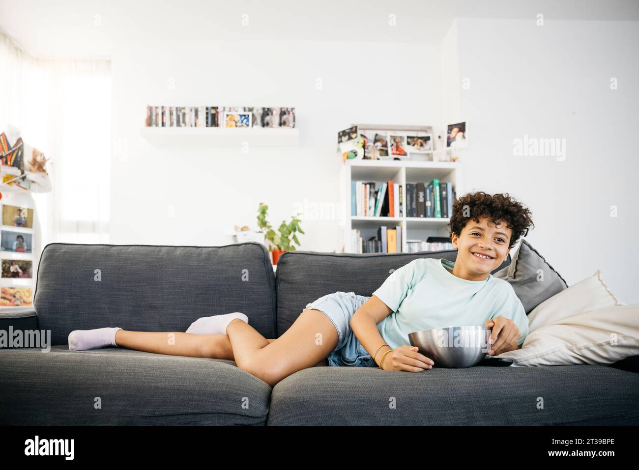 Fille ennuyée de dix ans allongée sur un canapé et regardant la télévision, avec une manette de télévision, dans un salon. Fille regardant un film à la maison. Banque D'Images