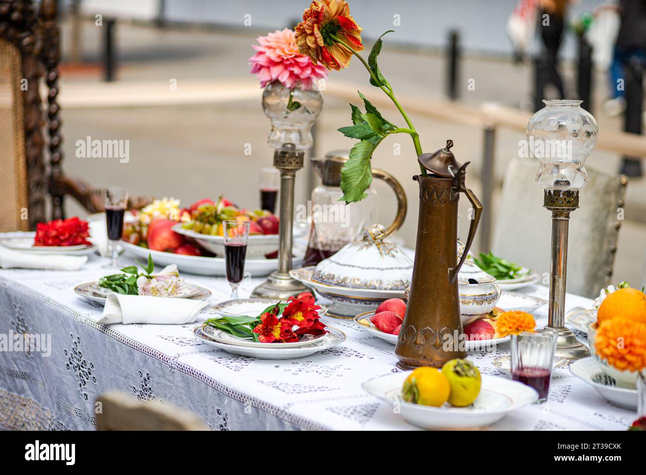 Une élégante table à manger extérieure ornée de fruits frais, de plats ornés, de lanternes vintage, d'un pichet en laiton et d'une fleur de dahlia vibrante sur une languette de dentelle Banque D'Images