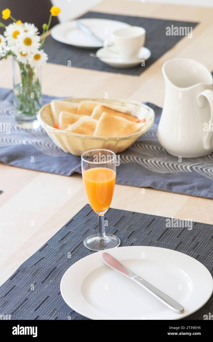 Un cadre de petit déjeuner confortable avec du jus d'orange frais dans un verre, des plats blancs, un pichet, et un panier de pain tissé, le tout accentué par des marguerites dans un vas Banque D'Images