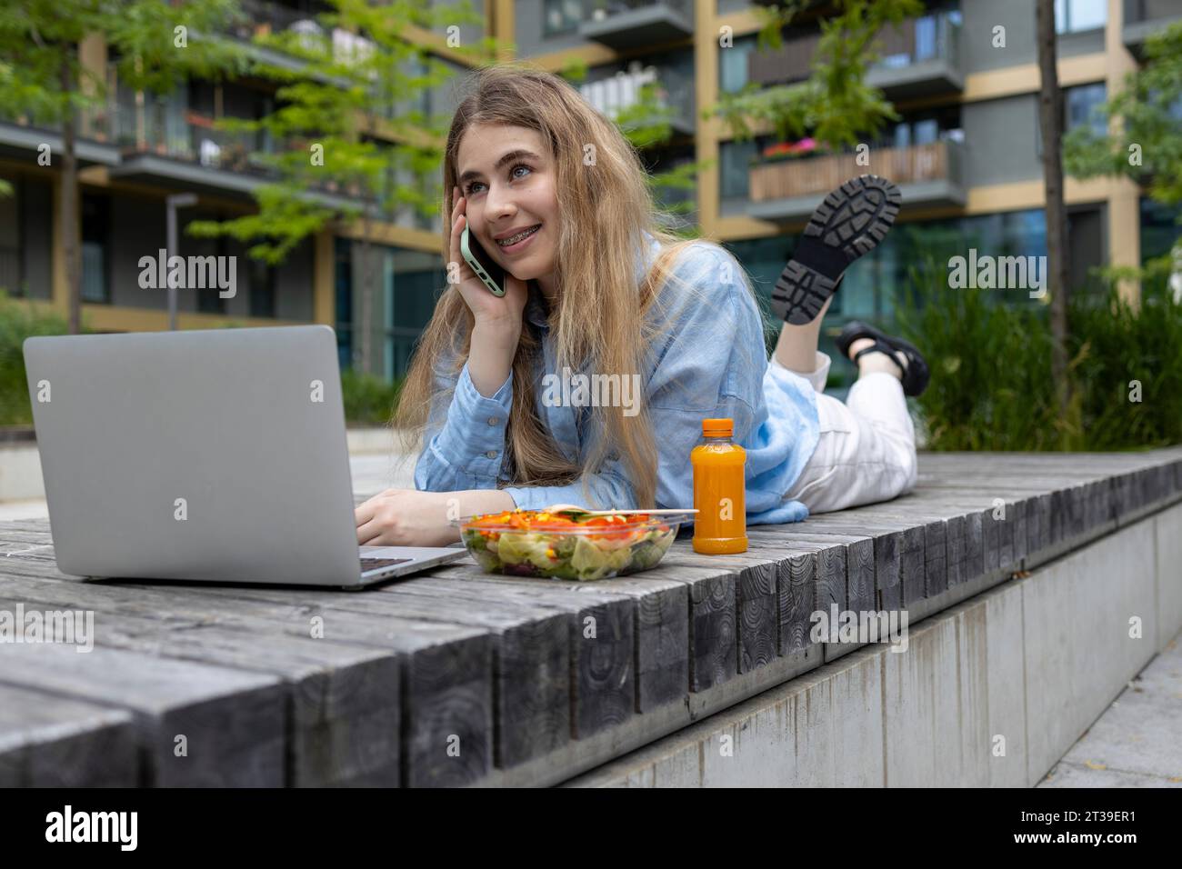 Heureuse femme étudiante aux cheveux blonds avec des bretelles couchées dans un banc en bois et parlant avec son smartphone à l'heure du déjeuner dans un lieu urbain. Banque D'Images