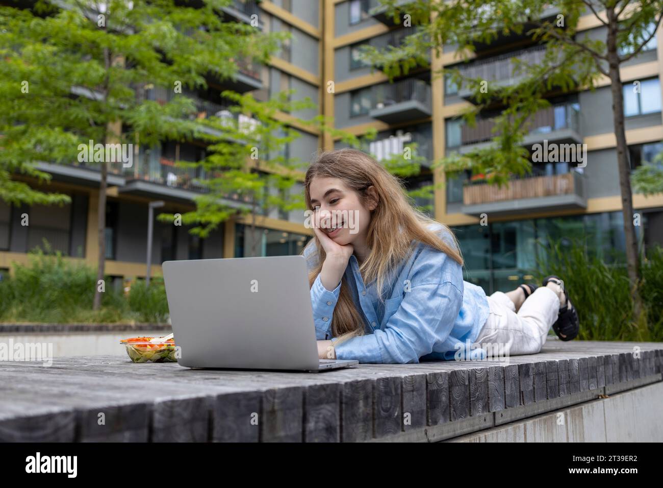 Heureuse femme étudiante aux cheveux blonds avec des bretelles couchées dans un banc en bois et regardant un ordinateur portable à l'heure du déjeuner à l'extérieur. Banque D'Images