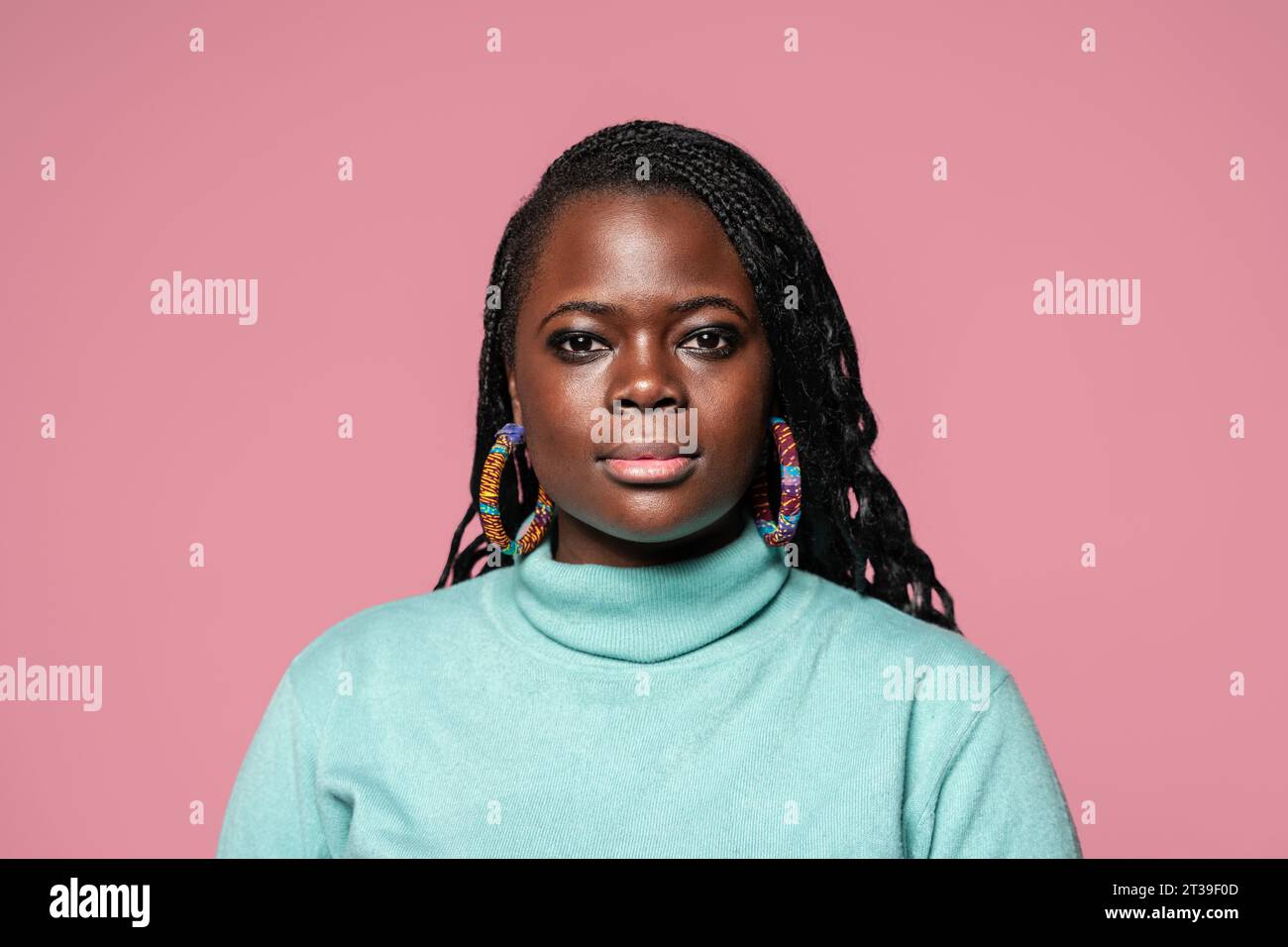 Portrait d'une femme africaine aux cheveux tressés portant des boucles d'oreilles colorées et un col roulé turquoise sur fond rose Banque D'Images