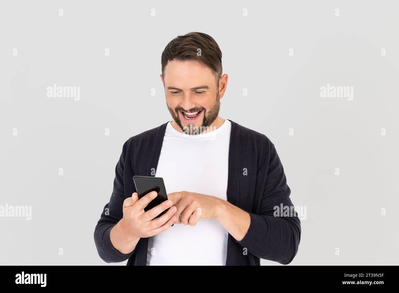 Heureux jeune homme barbu utilisant un téléphone intelligent mobile pour l'application mobile en ligne, les médias sociaux, sur fond gris. Ravi homme heureux exprimant surp Banque D'Images
