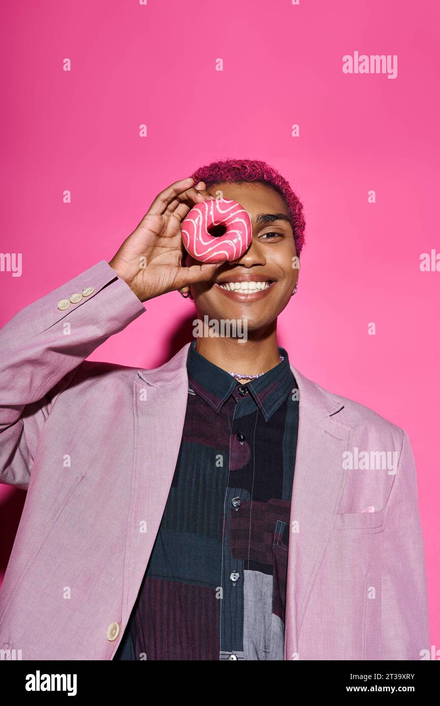 beau modèle masculin dans une tenue vibrante posant anormalement avec beignet près du visage sur fond rose Banque D'Images