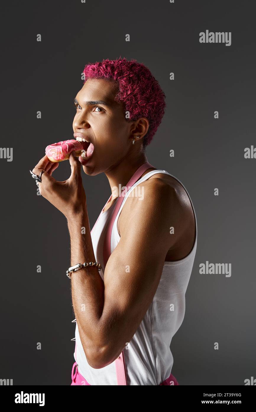 bon modèle masculin à la recherche avec des cheveux roses mangeant beignet posant sur fond gris, la mode et le style Banque D'Images