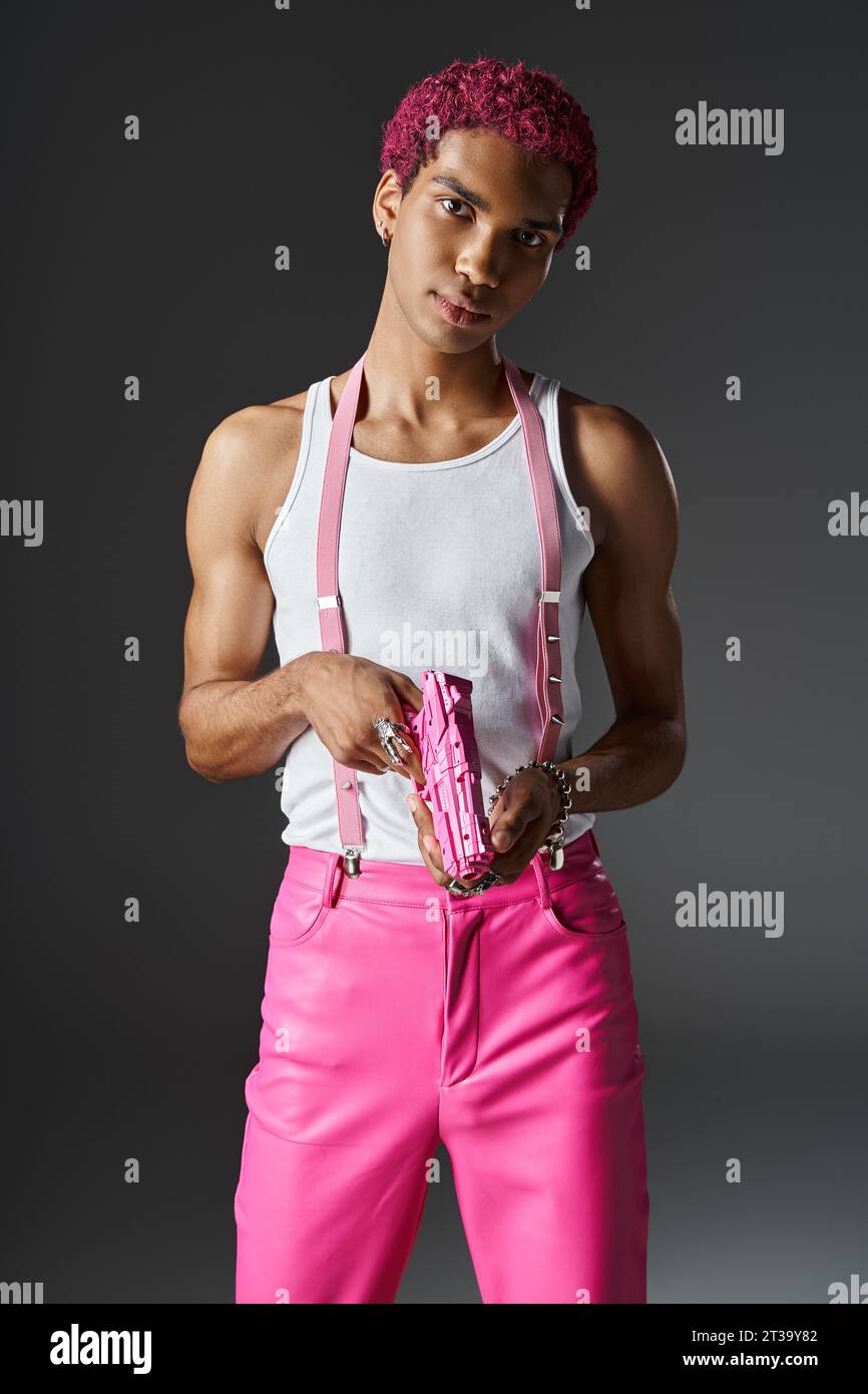 beau modèle masculin aux cheveux roses avec accessoires argentés et bretelles roses tenant pistolet jouet rose Banque D'Images