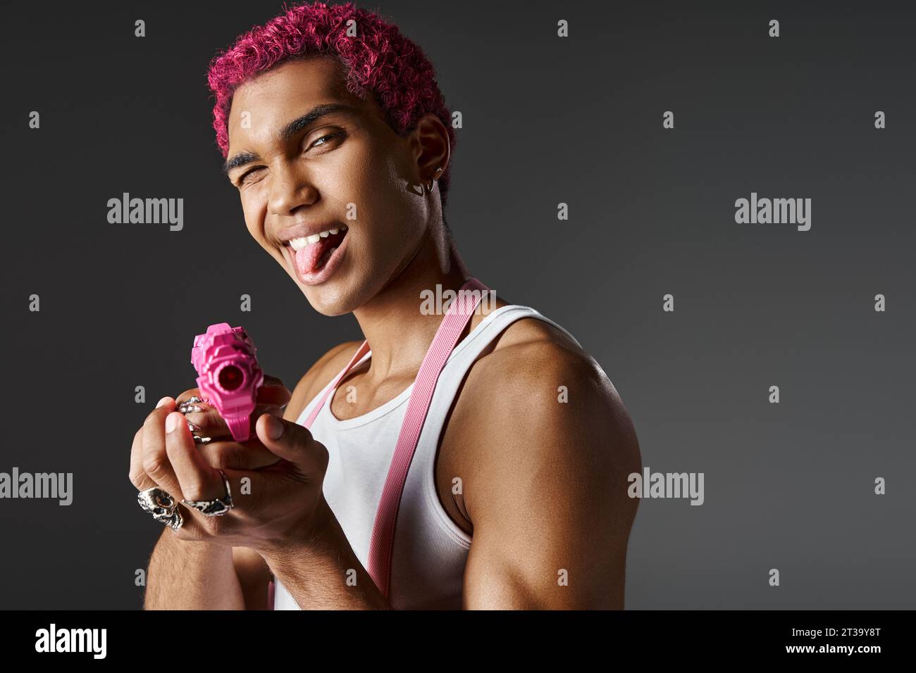 modèle masculin aux cheveux roses ludique sortant de sa langue et nivelant son pistolet jouet rose à la caméra Banque D'Images