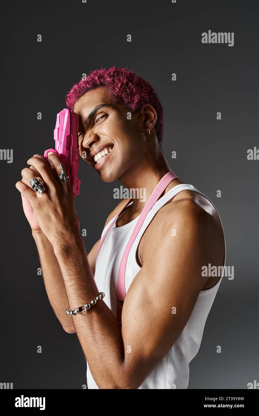 modèle masculin joyeux avec les cheveux roses bouclés posant dans le profil avec pistolet jouet rose et souriant à la caméra Banque D'Images