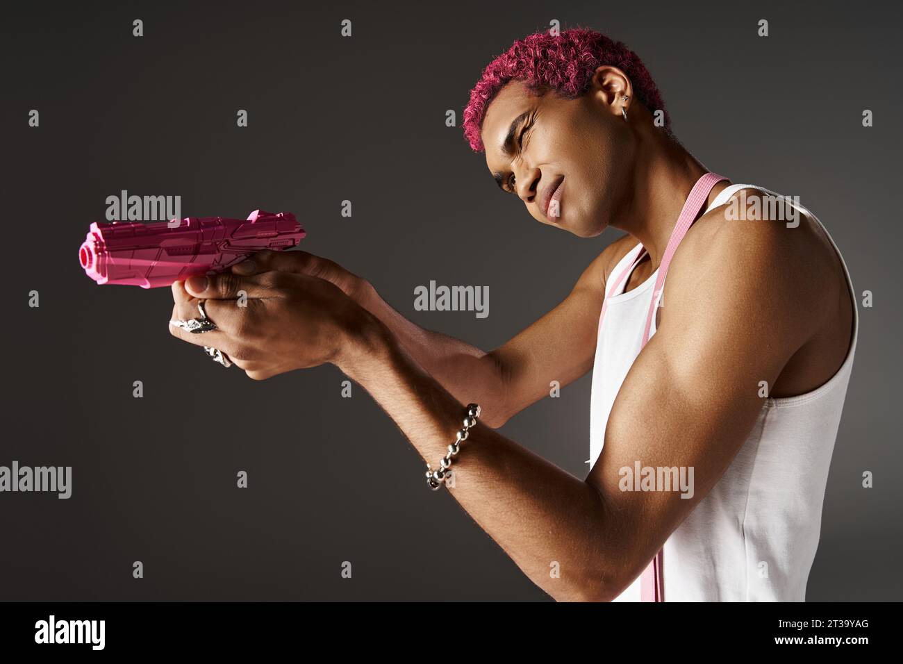 modèle masculin afro-américain aux cheveux roses en pantalon avec bretelles visant son pistolet jouet rose de côté Banque D'Images