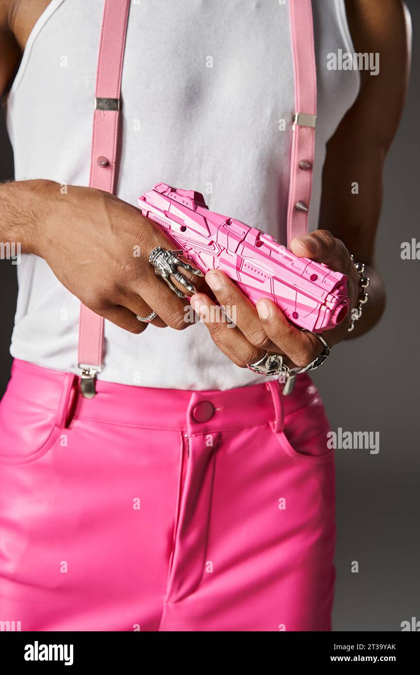 vue recadrée de jeune modèle masculin en pantalon rose et bretelles posant avec pistolet jouet sur fond gris Banque D'Images