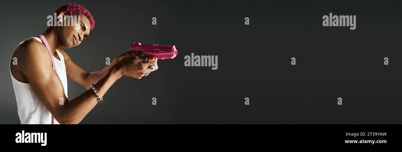 beau jeune modèle masculin dans une tenue vibrante avec des accessoires argentés visant son pistolet jouet rose loin Banque D'Images