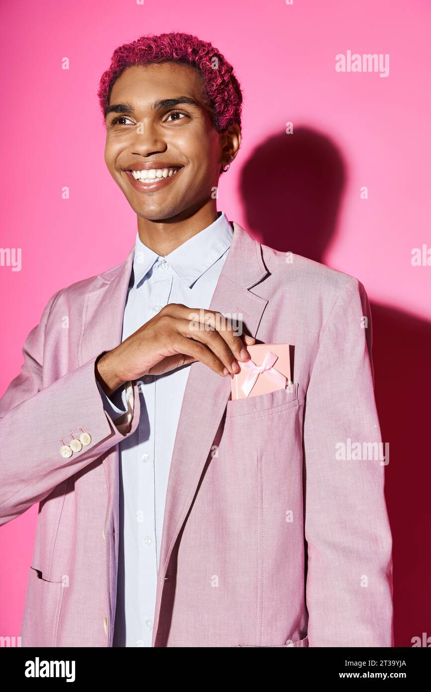 jeune modèle masculin aux cheveux roses dans des vêtements vibrants souriant anormalement avec présent dans sa poche Banque D'Images