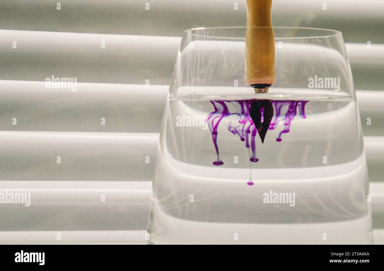 pointe métallique de stylo à encre trempée dans l'eau. diffusion de l'encre bleue dans une tasse en verre transparent avec de l'eau claire sur le fond d'un écran rayé Banque D'Images