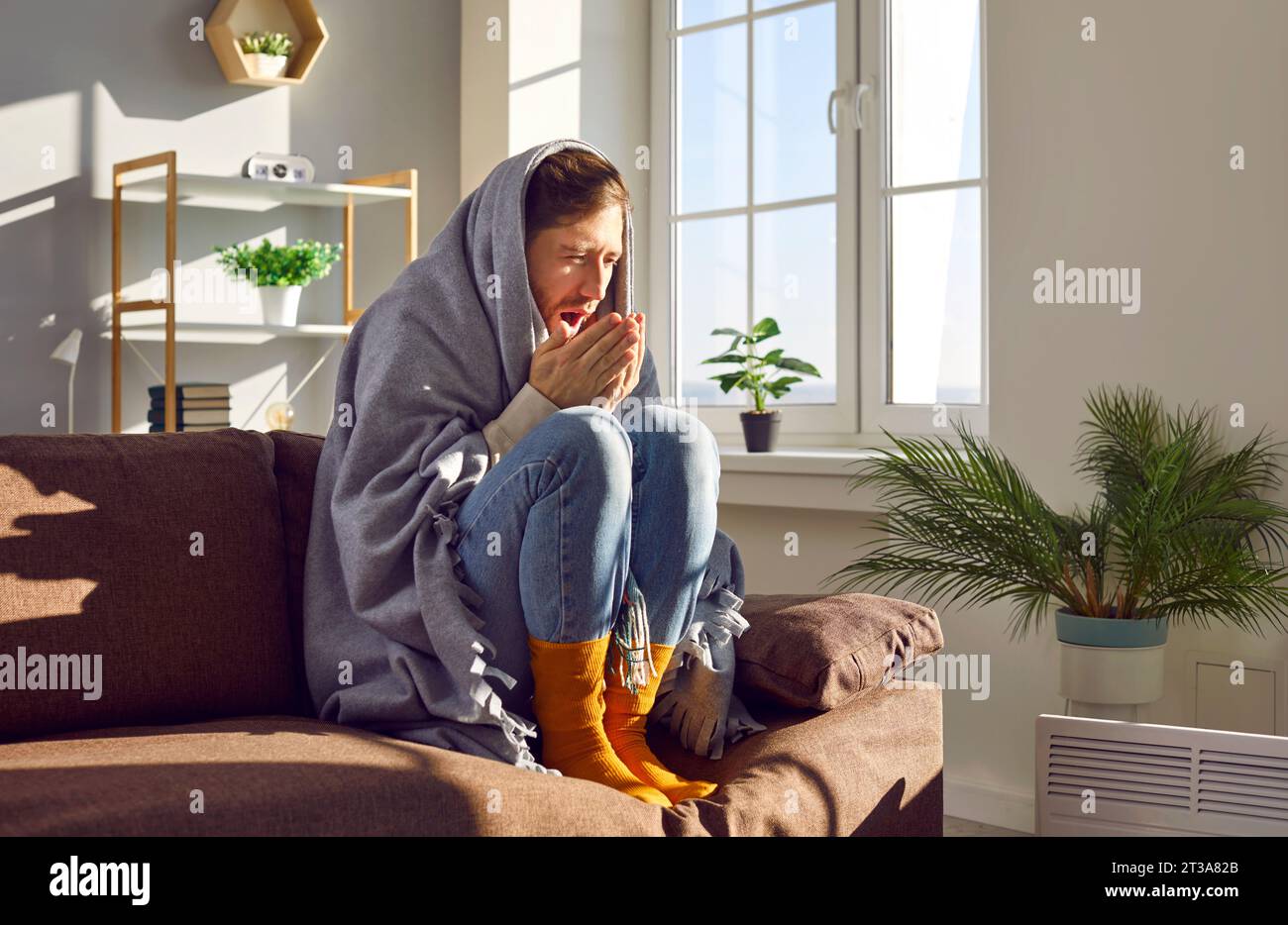 Couverture enveloppante pour jeune homme éternuant assis sur le canapé se réchauffant près de l'appareil de chauffage Banque D'Images