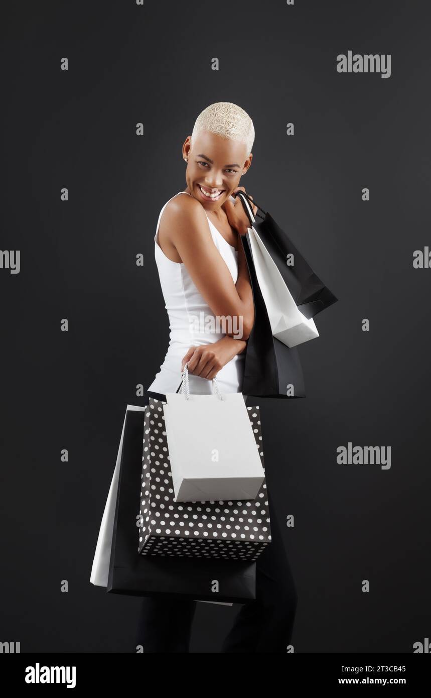 Black Friday shopping, une superbe femme noire souriante tenant de nombreux sacs noirs et blancs, sur un fond noir, regarde dans l'appareil photo, heureux de h Banque D'Images