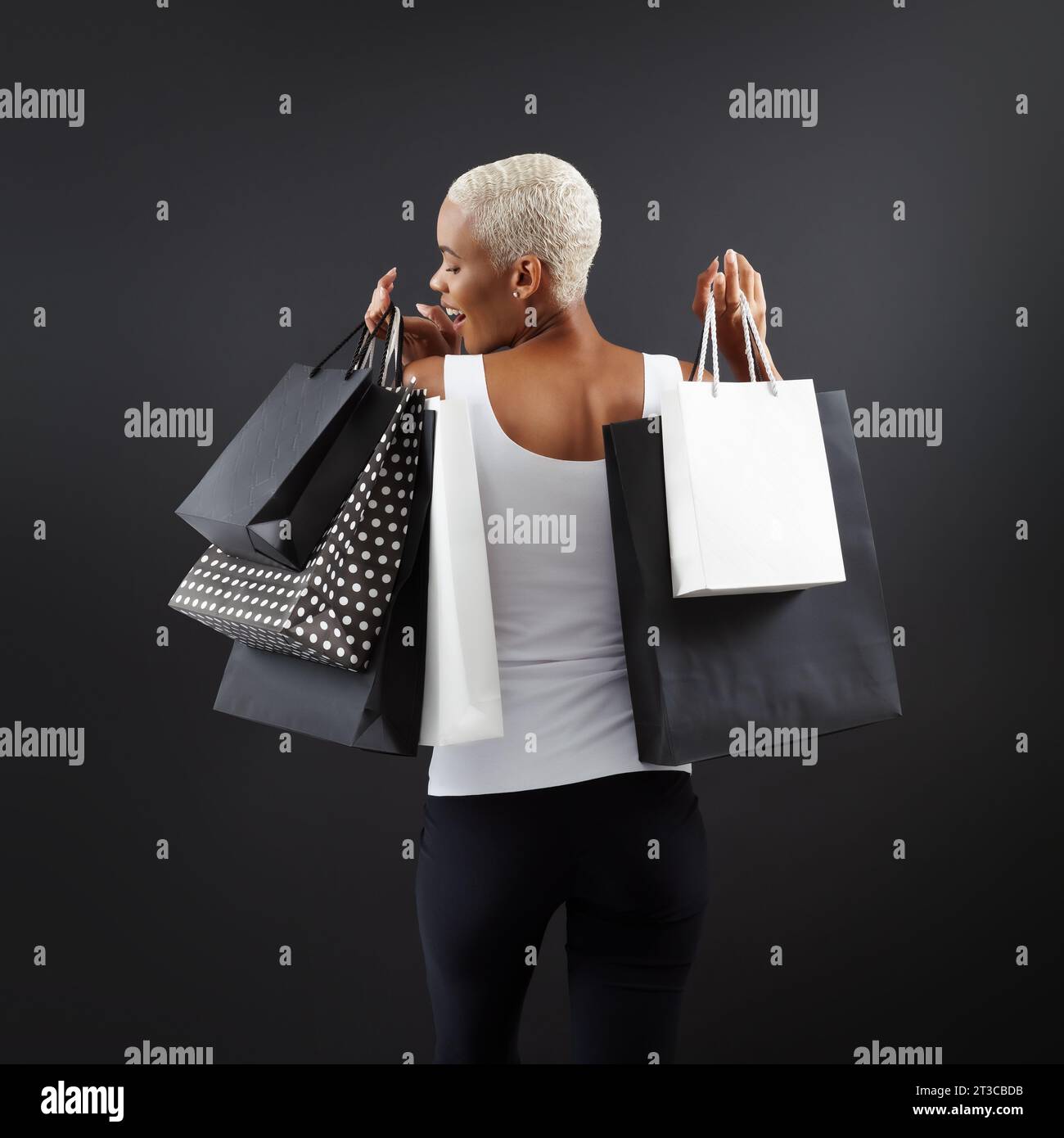 Black Friday shopping, superbe femme noire souriante tenant de nombreux sacs noirs et blancs, sur un fond noir, vue de derrière, heureuse d'avoir trouvé TH Banque D'Images