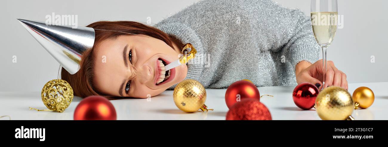 Bannière de femme dans la casquette de fête couchée sur la table près du champagne et boules de Noël, soufflant corne de fête Banque D'Images