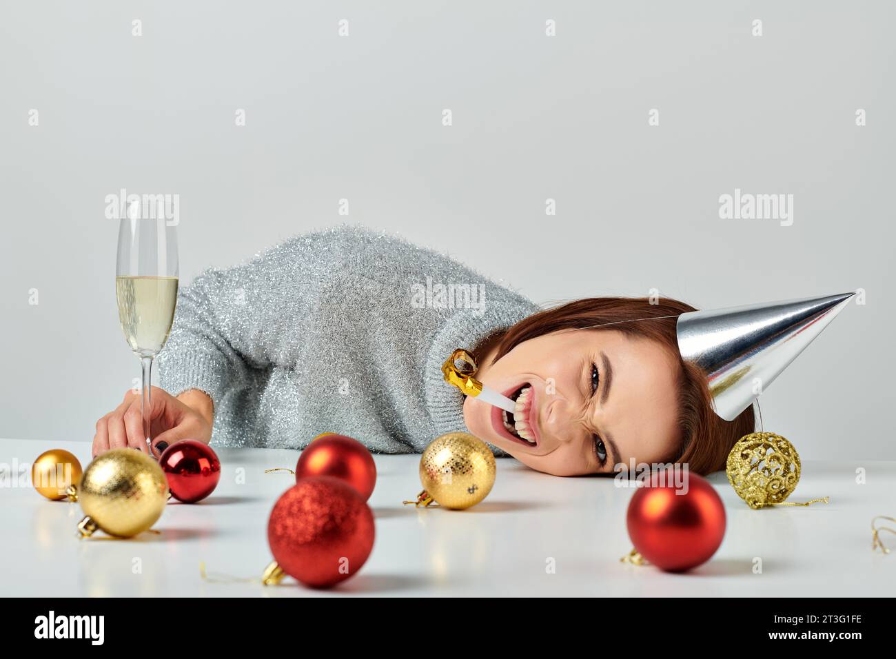 Femme dans la casquette de fête couchée sur la table près du verre de champagne et des boules de Noël, soufflant corne de fête Banque D'Images