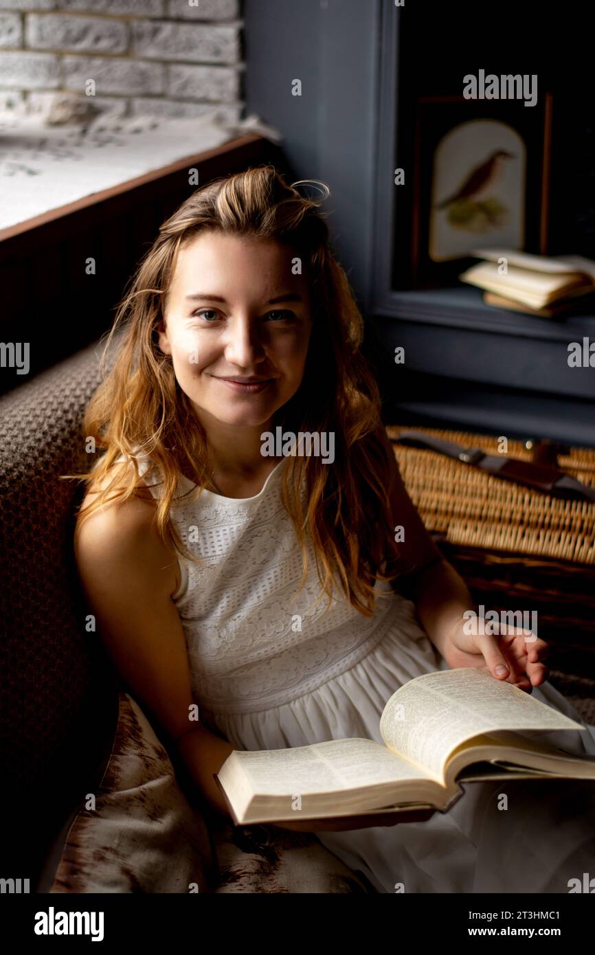 Assise sur le canapé, une jeune femme à l'apparence attrayante et aux cheveux longs sourit chaleureusement tout en tenant un livre. La photo parle du plaisir Banque D'Images