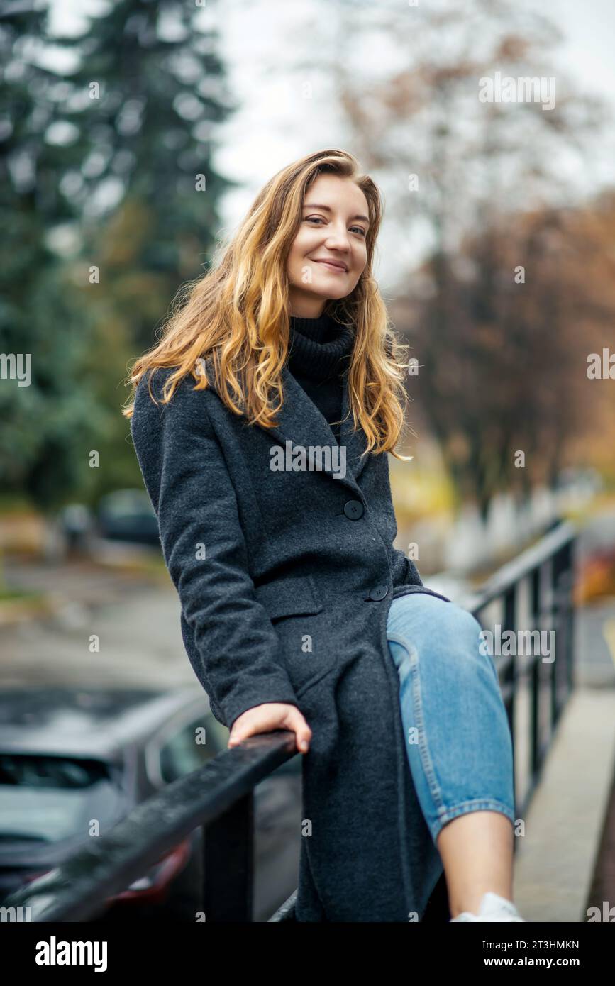 Une femme dans un long manteau gris et un Jean bleu sur des baskets blanches est assise sur la rampe et regarde la caméra dans la rue d'une ville d'automne Banque D'Images
