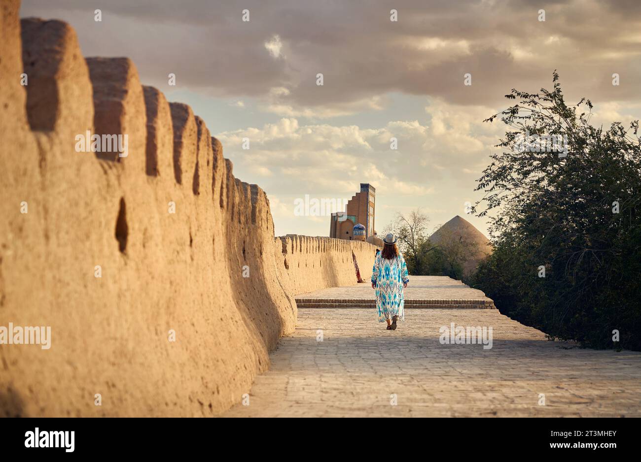 Jeune femme en robe ethnique avec ornement bleu marchant au mur de la ville dans Itchan Kala ancienne ville de Khiva en Ouzbékistan. Banque D'Images