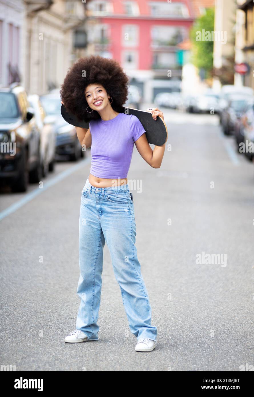 Corps entier de jeune femme afro-américaine positive avec les cheveux bouclés posant avec skateboard sur la rue de la ville Banque D'Images