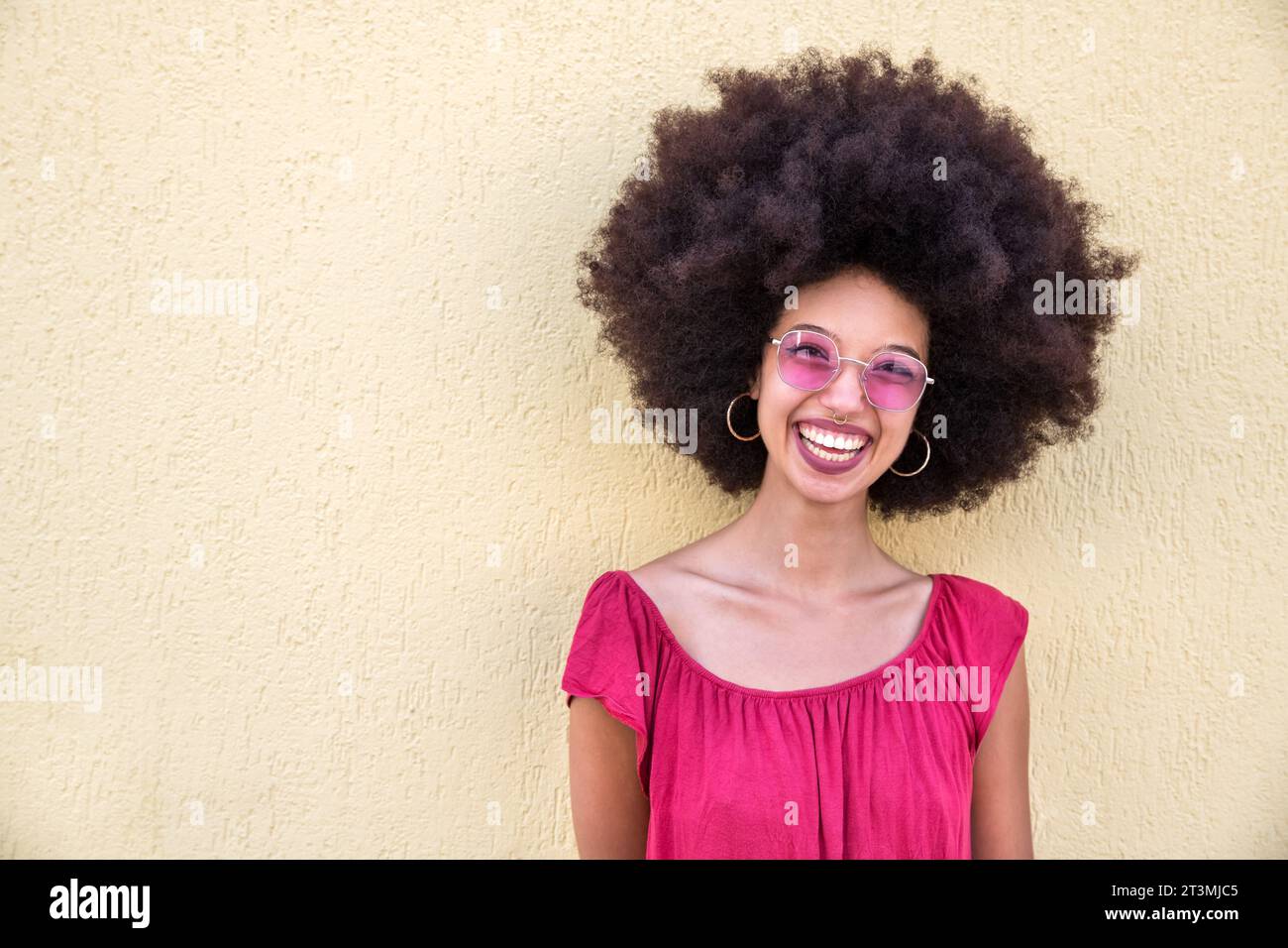 Portrait de jeune femme marocaine heureuse avec coiffure afro et lunettes de soleil élégantes debout près du mur beige et regardant la caméra Banque D'Images