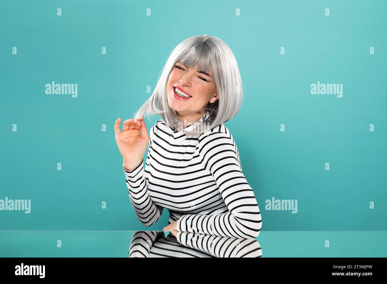 Positif jeune modèle féminin en chemise rayée à manches longues touchant les cheveux gris et fermant les yeux tout en étant assis à la table miroir en studio Banque D'Images