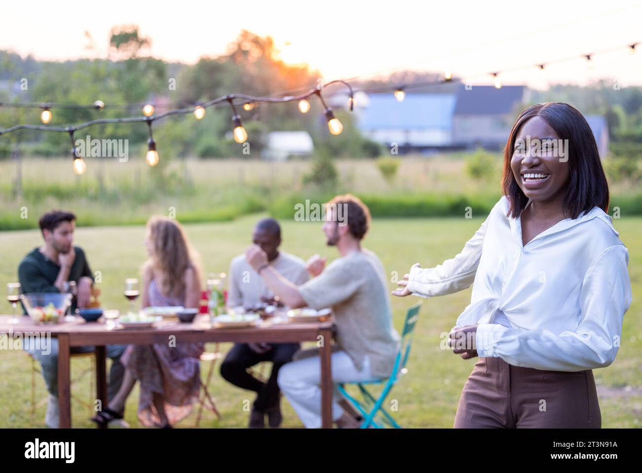 Un rassemblement joyeux par une journée ensoleillée encapsule une belle jeune femme noire dans un chemisier blanc, riant et savourant un barbecue de jardin parmi les amis, le vin et le plaisir de pique-nique en plein air. L'essence de la camaraderie et des festivités estivales magnifiquement capturées en haute résolution. Radiant Jeune femme appréciant Sunny Garden BBQ avec des amis. Photo de haute qualité Banque D'Images