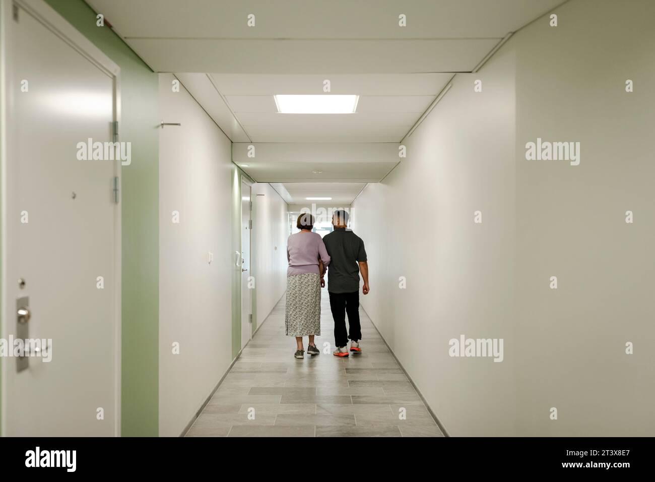 Vue arrière sur toute la longueur de la femme âgée marchant bras dans bras avec l'infirmière masculine dans le couloir de la maison de retraite Banque D'Images