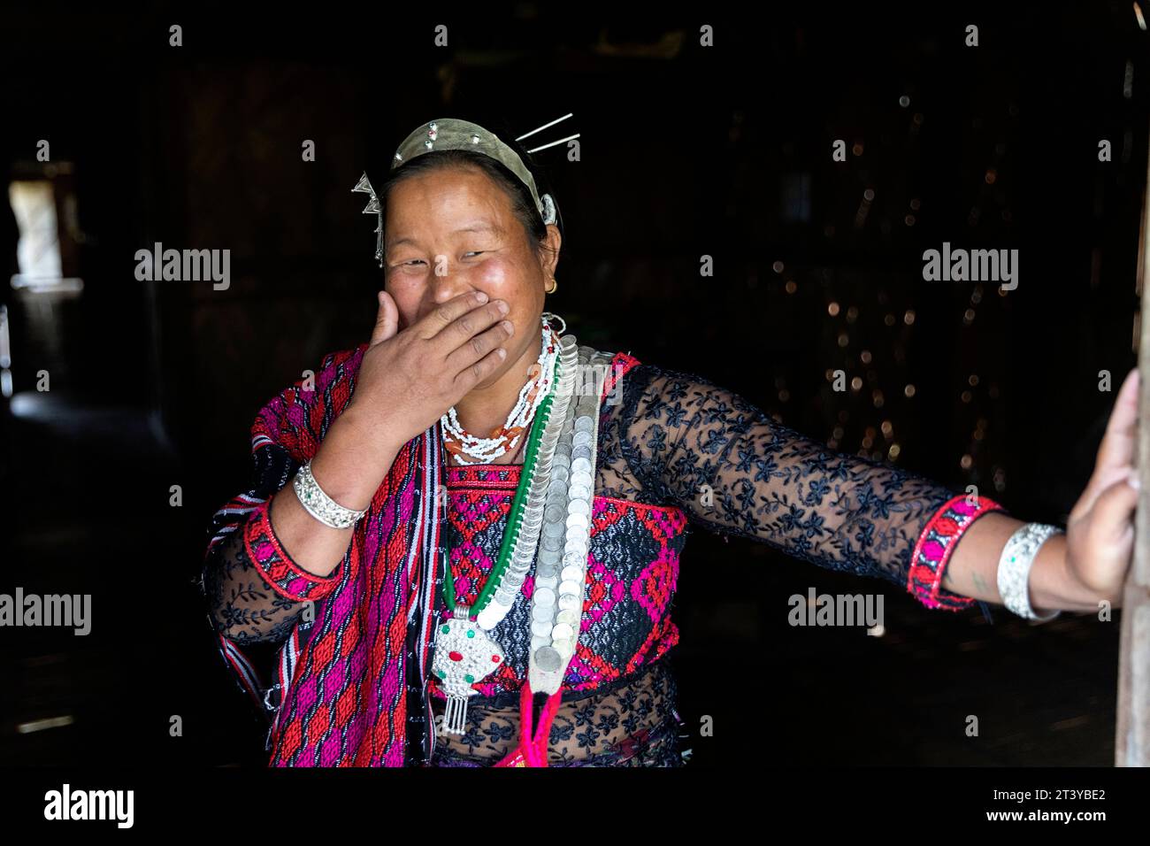 Femme de la tribu Adi en vêtements traditionnels souriant avec la main sur la bouche, appuyée sur la porte ouverte dans une maison traditionnelle en bois à Assam dans le nord-est de l'Inde Banque D'Images