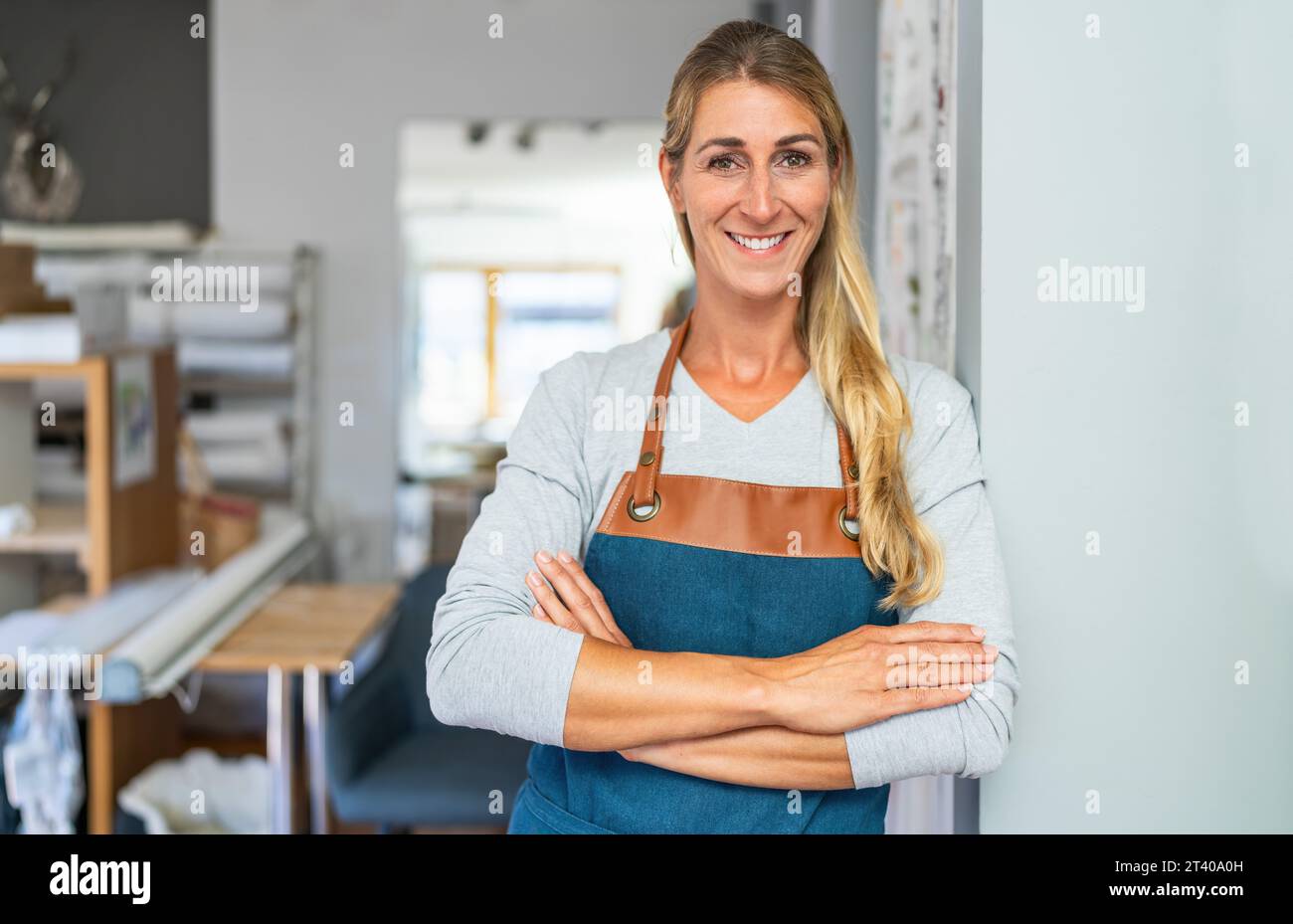 femme blonde joyeuse avec les bras croisés en tablier se tient dans un espace de travail de tailleur. Elle a un ruban à mesurer et des ciseaux dans sa poche de tablier et est sur Banque D'Images