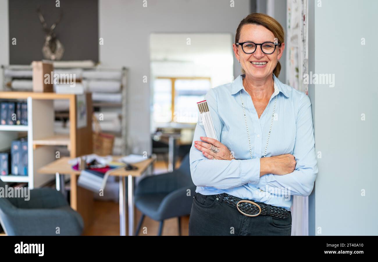 Femme tailleur souriante professionnelle avec une tresse de cheveux, portant des lunettes et une chemise rayée bleue, se tient dans un espace de travail moderne et tenant une règle. Banque D'Images