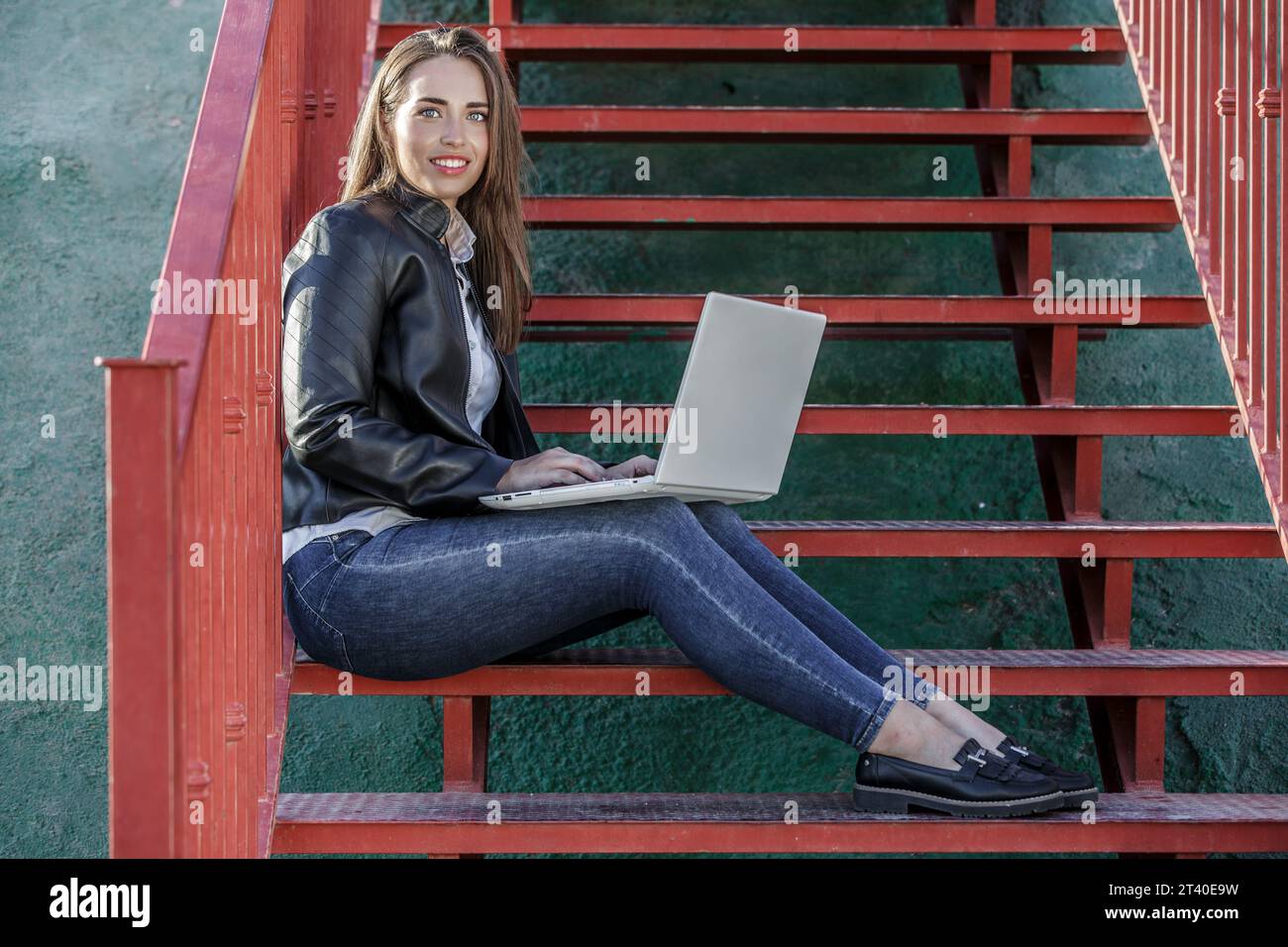 Corps entier de jeune femme heureuse travailleur à distance assis sur l'escalier rouge et travaillant à distance via netbook par jour ensoleillé Banque D'Images
