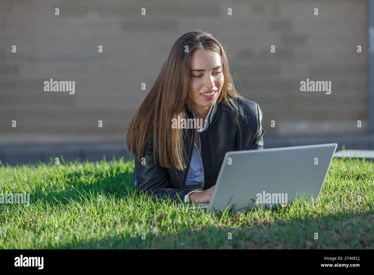Souriant jeune femme travailleur à distance dans la veste en cuir couché sur la pelouse tout en tapant du texte sur netbook travaillant en ligne sur le projet à l'extérieur Banque D'Images