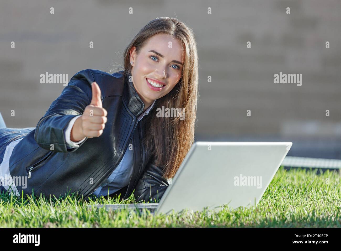 Satisfait jeune femme travailleur à distance dans la veste en cuir couché sur l'herbe à l'aide d'un ordinateur portable tout en montrant le geste du pouce vers le haut pendant le travail en ligne à l'extérieur Banque D'Images