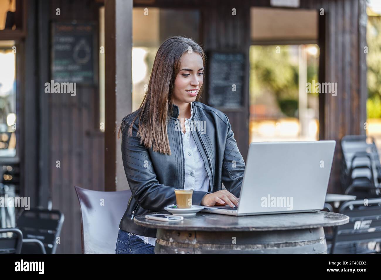 Positive jeune femme freelance dans la veste en cuir noir assis à la table avec tasse à café et la navigation netbook tout en travaillant sur le projet à distance dans outd Banque D'Images