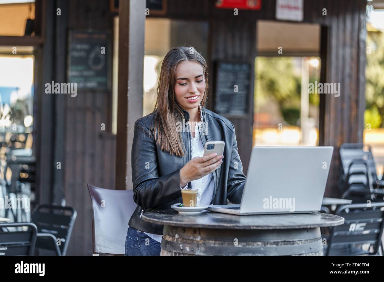 Positive jeune femme entrepreneur en tenue décontractée assis à la table ronde avec netbook et tasse de café chaud tout en discutant sur téléphone portable pendant remot Banque D'Images