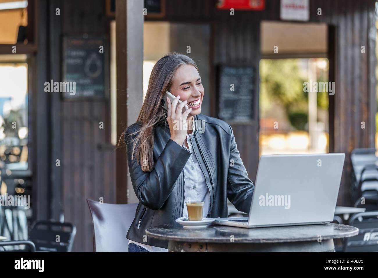 Heureuse jeune femme dans la veste en cuir assis contre le café flou tout en naviguant sur ordinateur portable et ayant une conversation agréable sur téléphone portable pendant la télécommande Banque D'Images