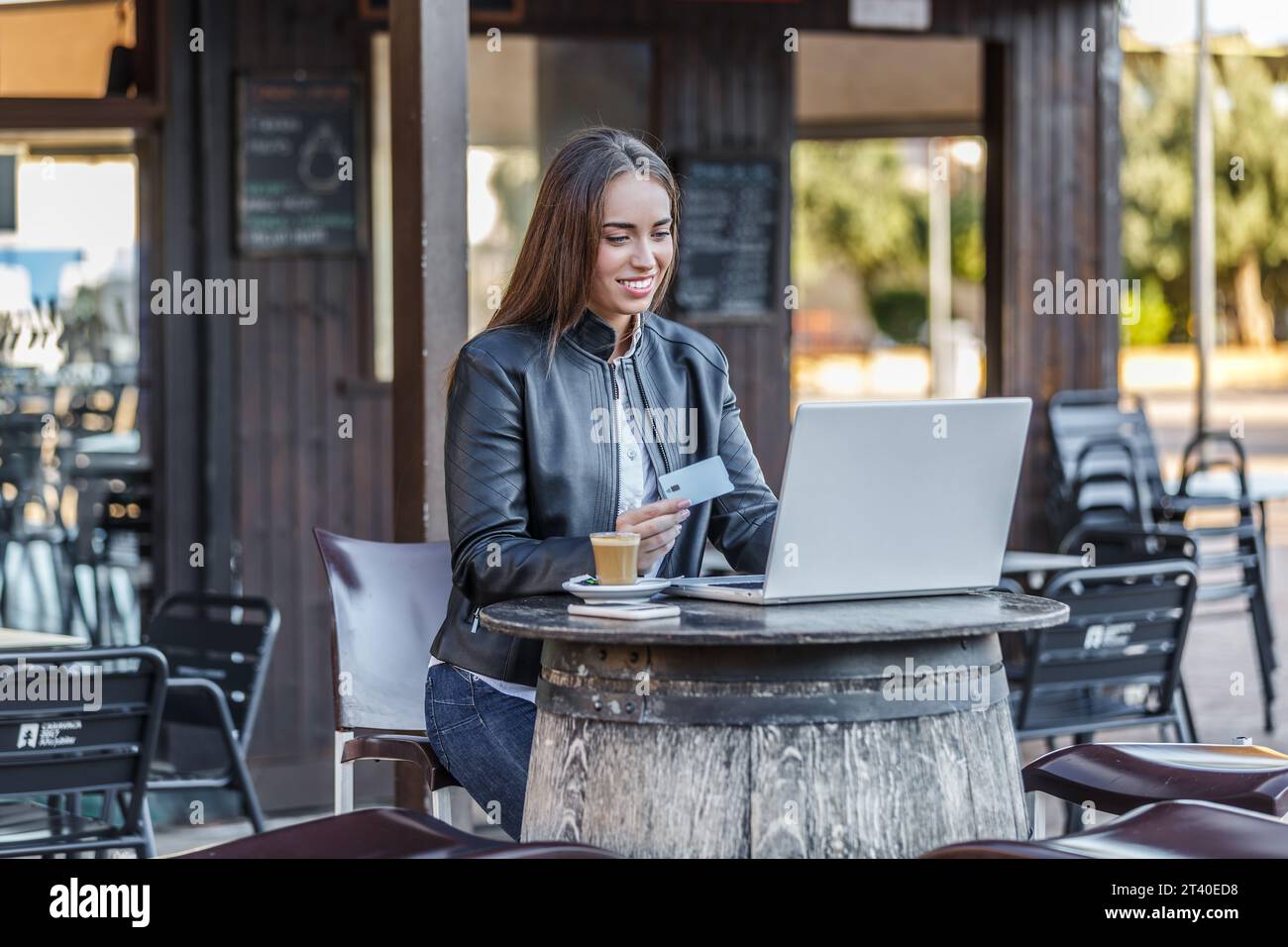 Jeune femme souriante en tenue décontractée assise à la table ronde dans la cafétéria de rue avec tasse de café tout en utilisant un ordinateur portable et en payant pour les achats en ligne Banque D'Images