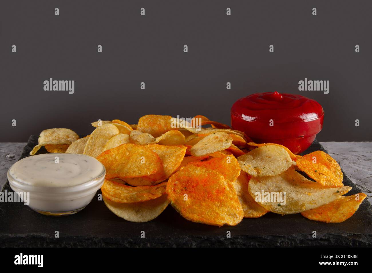 Vue latérale du tas de chips de pommes de terre avec deux sauces trempées sur la planche de service en ardoise noire sur la table en plâtre gris. Assortiment savoureux de trempettes Banque D'Images