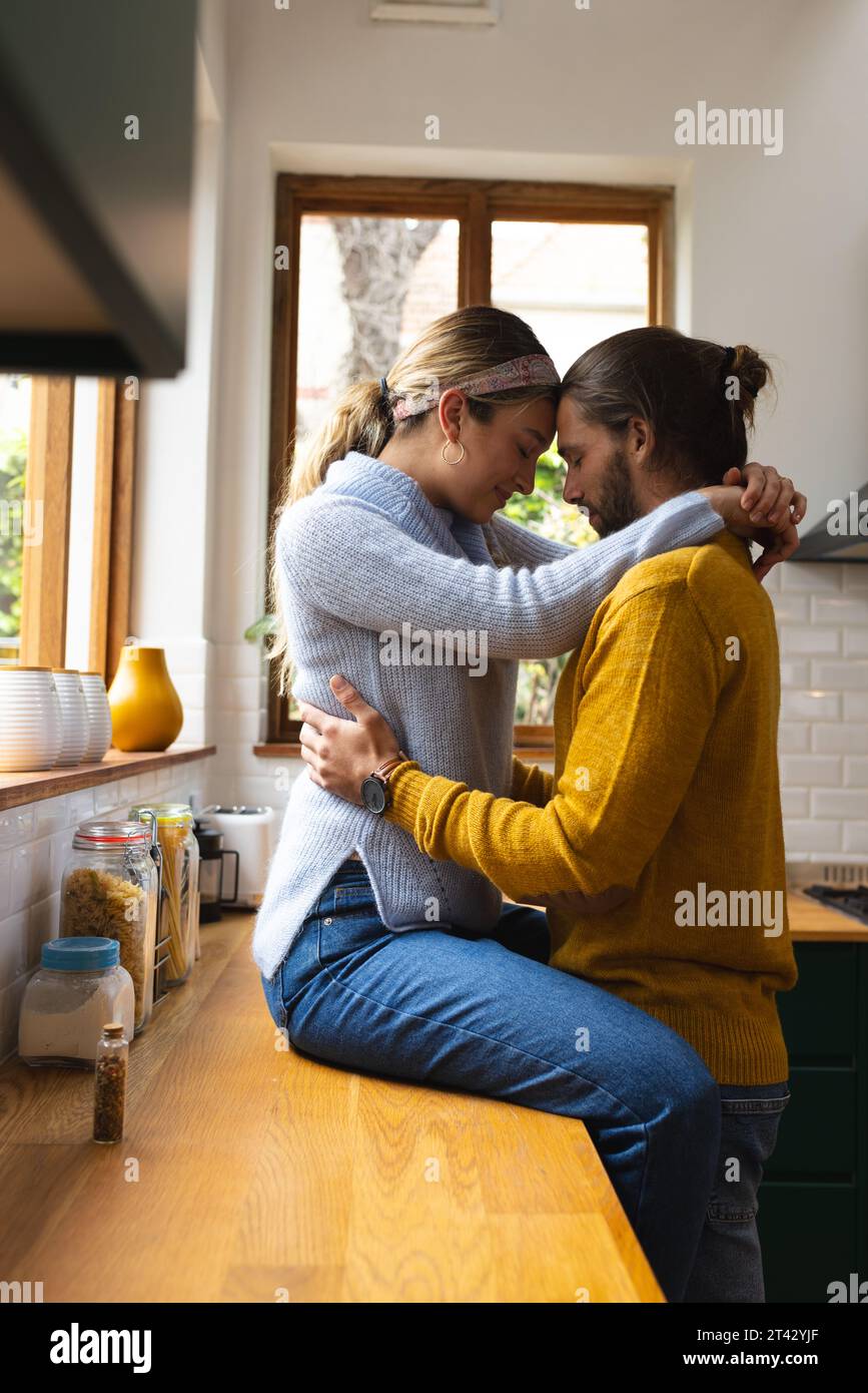 Heureux, romantique couple caucasien embrassant assis sur le plan de travail et debout dans la cuisine, espace de copie Banque D'Images